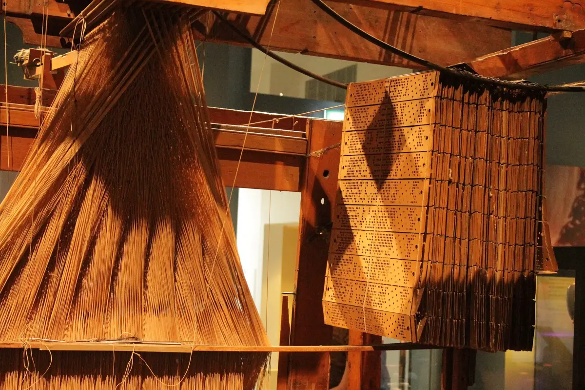 A wooden Jacquard loom with its chain of punched cards hanging beside thousands of threads.